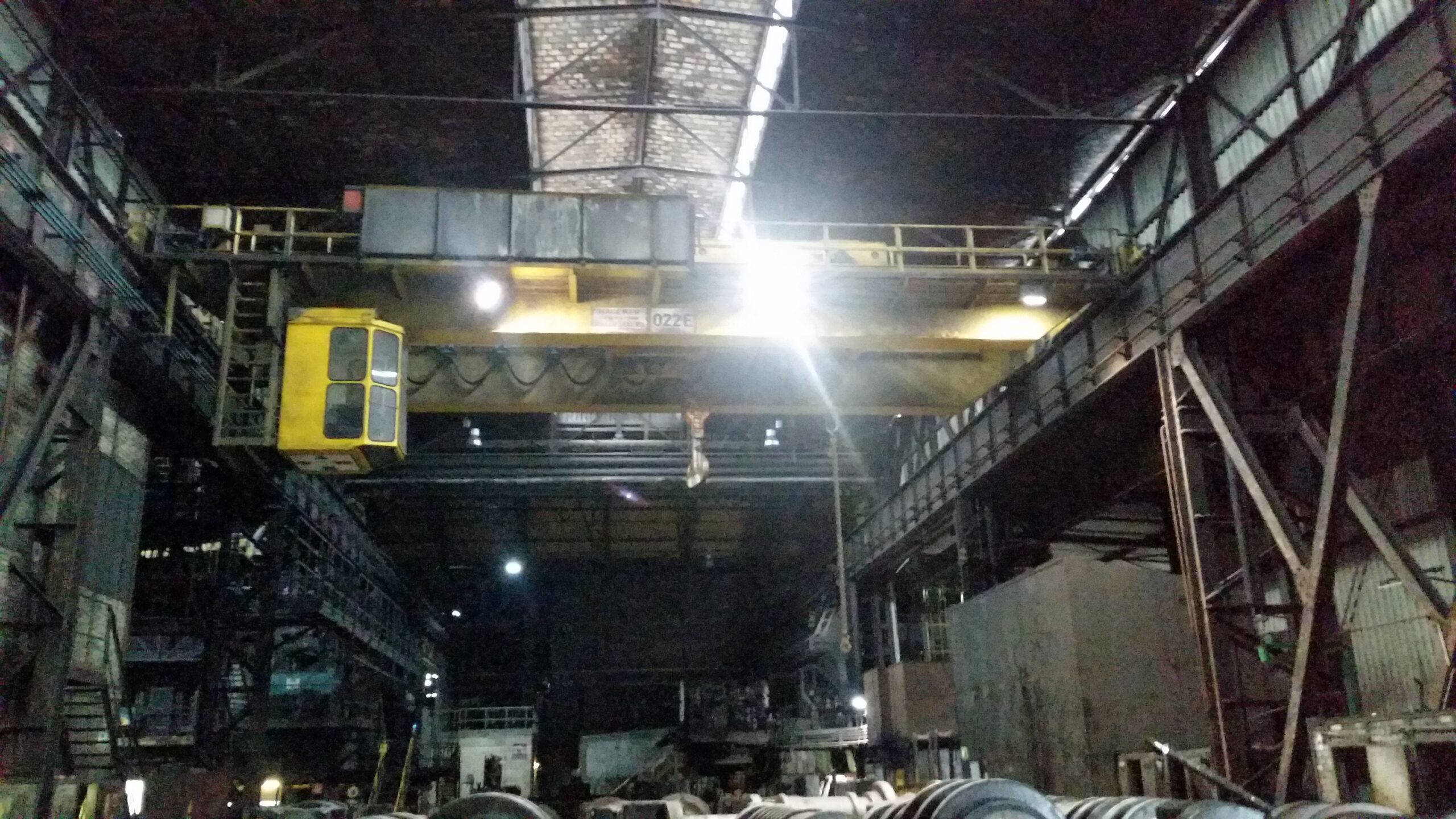 Im Inneren der Produktionshalle ist ein Brückenkran mit Steuerkabine zu sehen, der unter der Decke inmitten von Metallkonstruktionen und Maschinen angebracht ist.