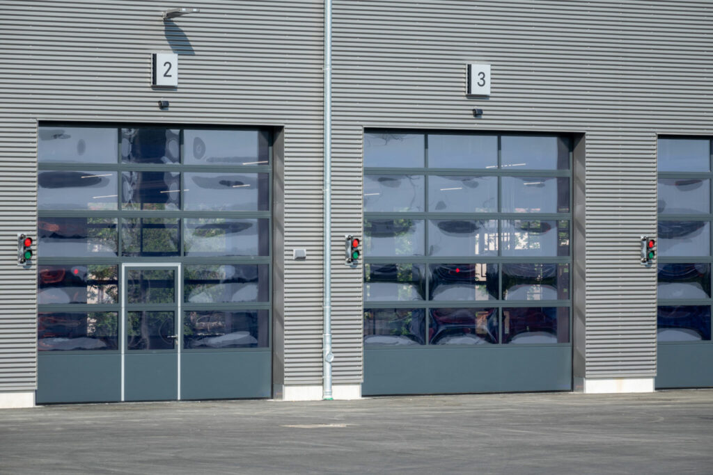 Die Fassade eines Industriegebäudes mit mehreren Sektionaltoren und Glaselementen; daneben sind Signalleuchten angebracht, die den Betrieb der Einfahrtssysteme anzeigen.