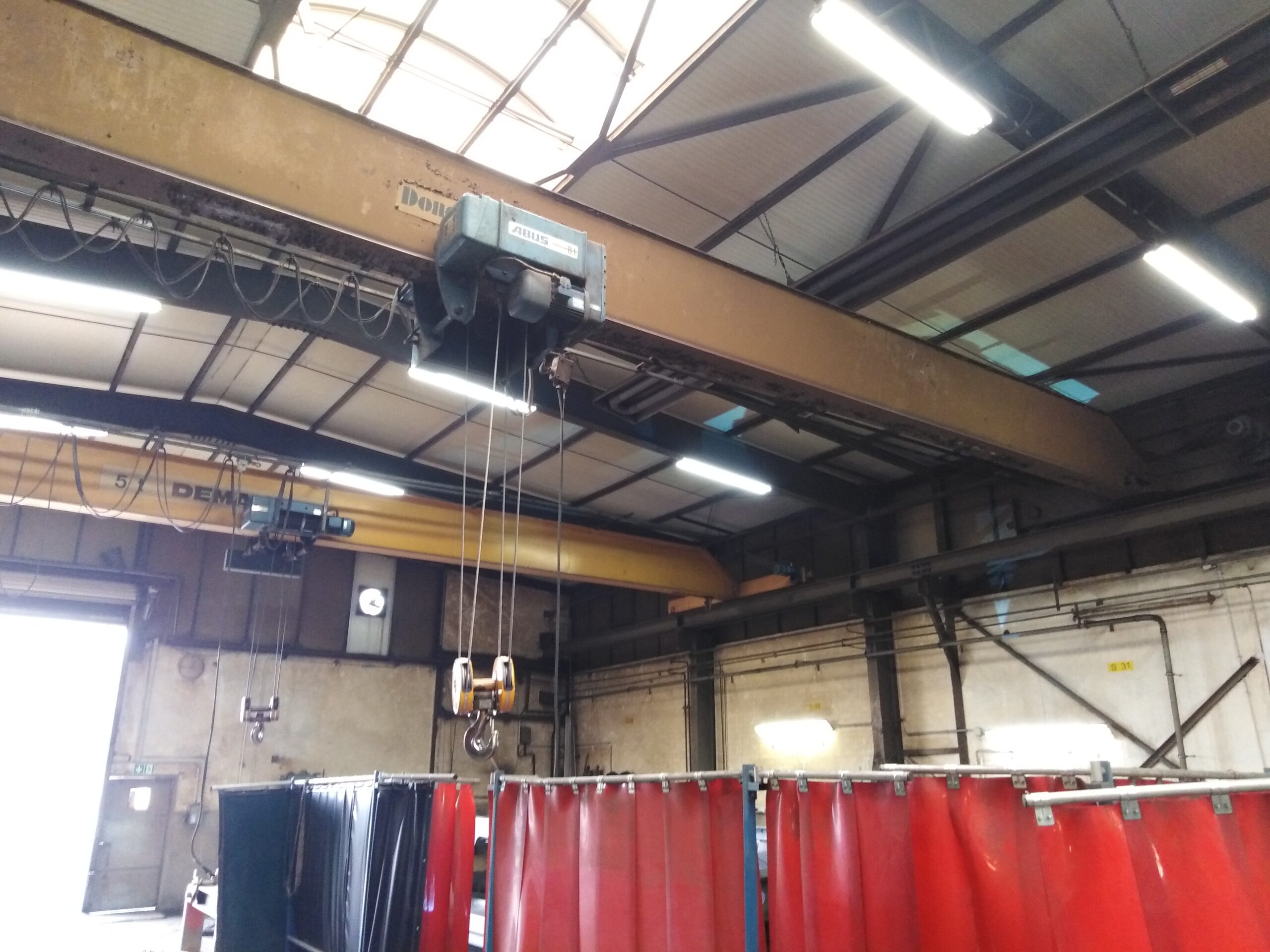 In der Produktionshalle ist an der Decke ein Kran mit Hebevorrichtung und Haken installiert; darunter befinden sich die Arbeitsbereiche, die durch Schutzvorhänge abgetrennt sind.