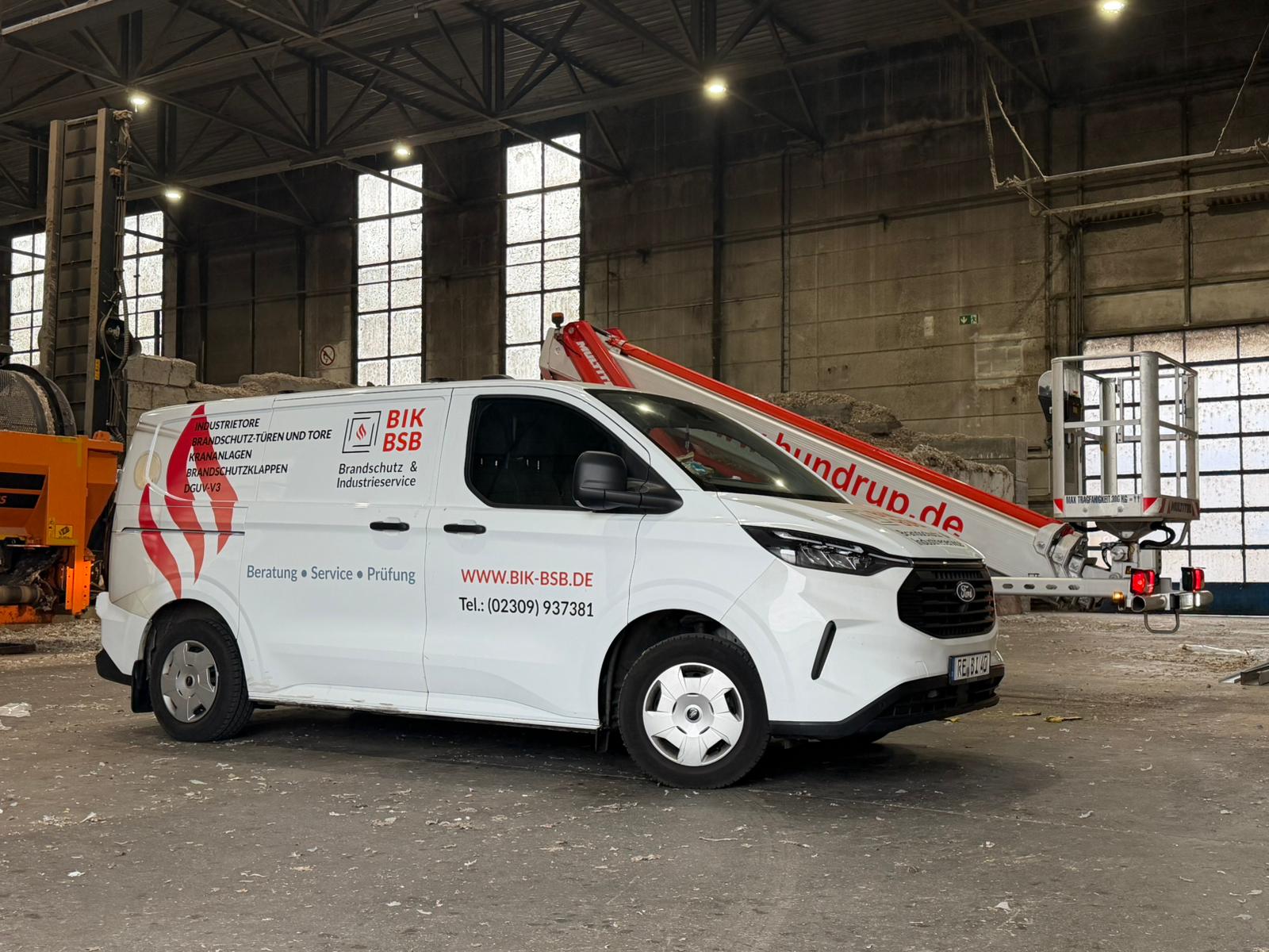 Ein weißer Servicewagen mit dem Logo von BIK-BSB und einer Hebebühne steht in einer Industriehalle mit hoher Decke und großen Fenstern