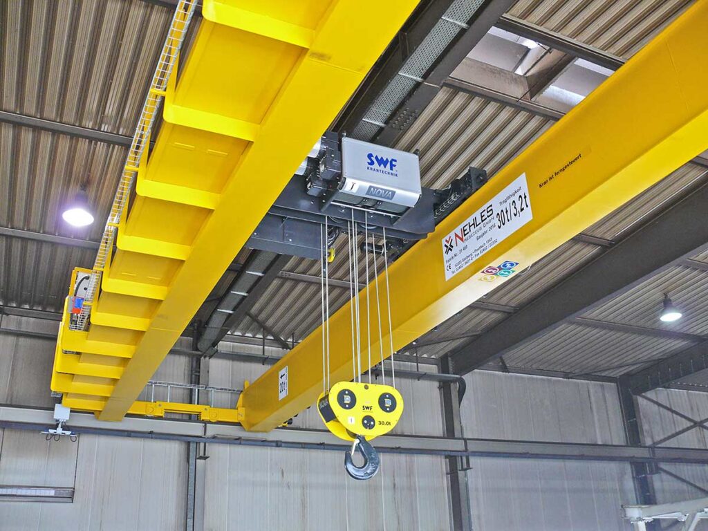Ein gelber Kranbalken mit elektrischem Hubwerk und Haken, der an der Decke einer Industriehalle angebracht ist.