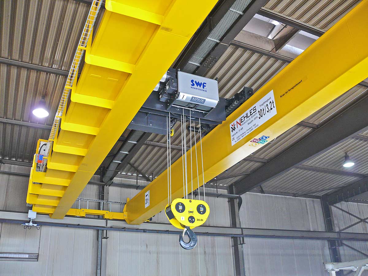 Ein gelber Kranbalken mit elektrischem Hubwerk und Haken, der an der Decke einer Industriehalle angebracht ist.